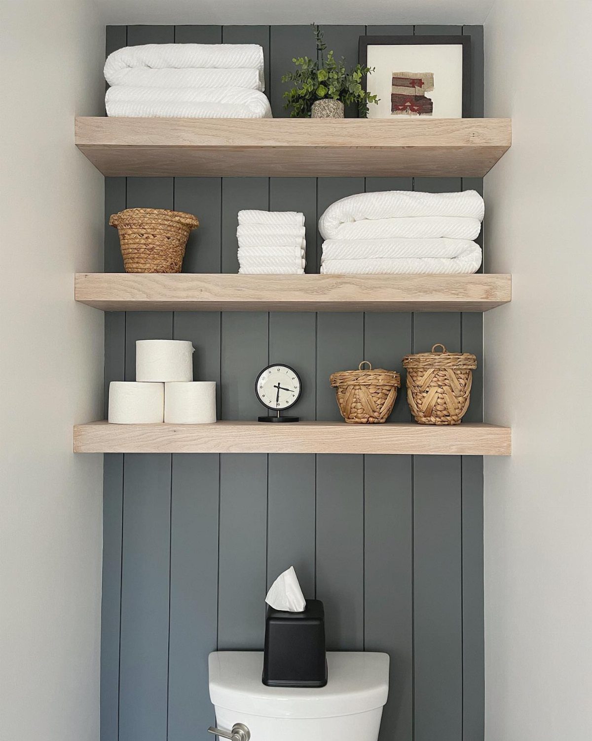 Three wooden floating shelves mounted on a grey paneled wall above a white toilet.