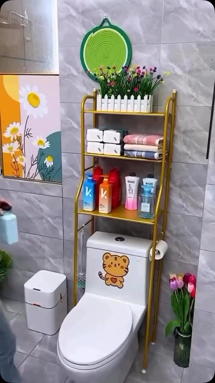Gold metal tiered shelving unit holding flowers, towels, and toiletries standing behind a white toilet.