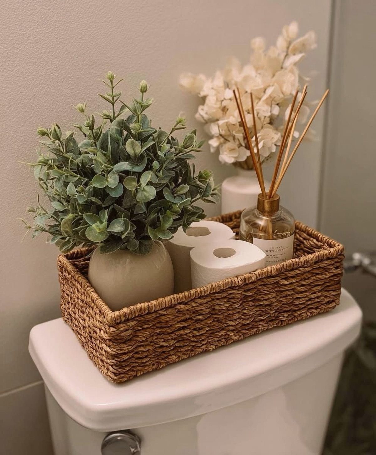 A woven basket containing a potted plant, toilet paper rolls, and a reed diffuser on a toilet tank.