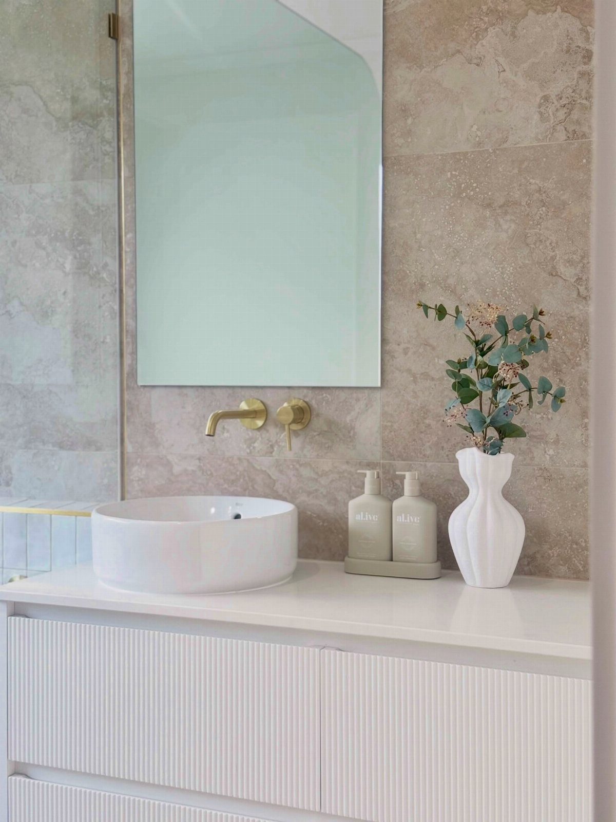 White bathroom vanity with a vessel sink, gold fixtures, and a large frameless mirror.