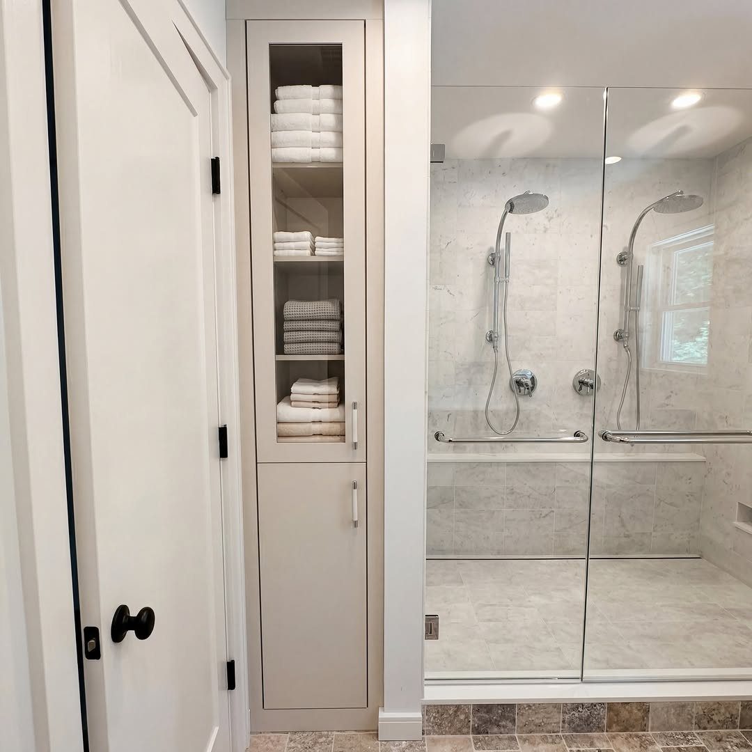 A tall white linen tower cabinet with glass doors filled with folded white towels next to a shower.