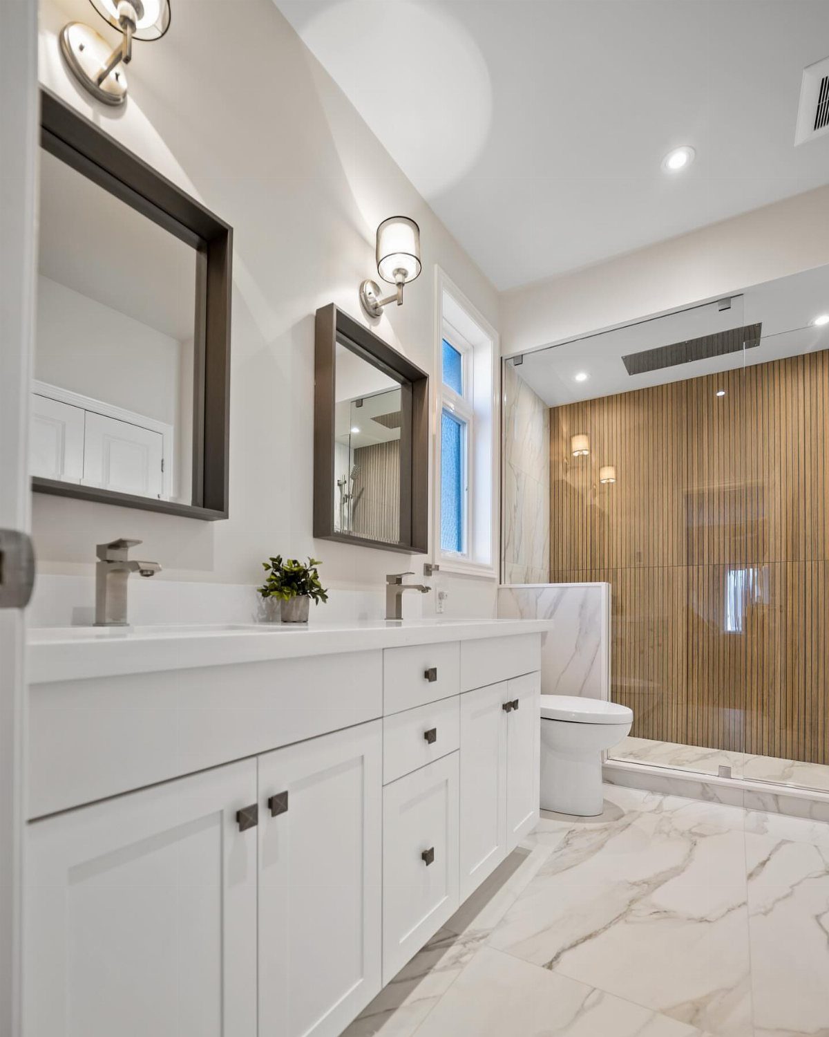 White bathroom vanity with shaker cabinets, two mirrors, and marble tile flooring in a bright room.