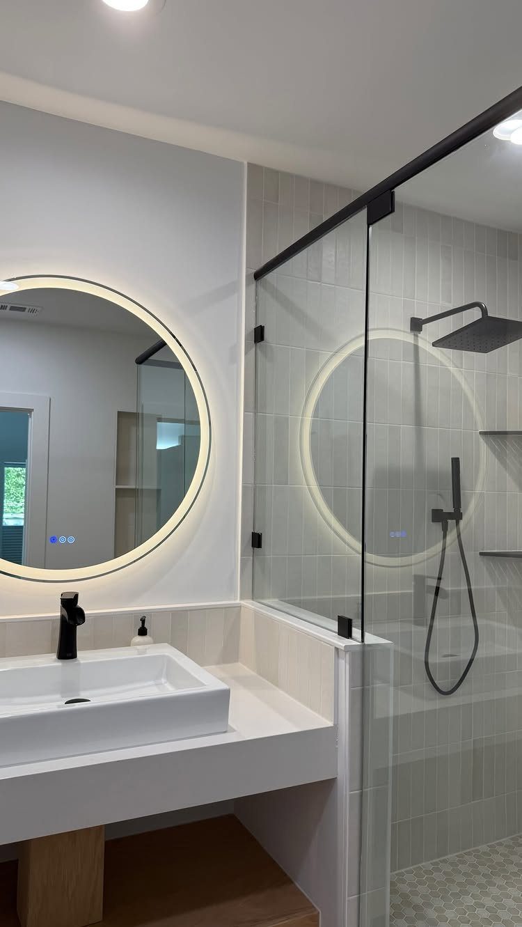 Modern bathroom with a white vessel sink, circular backlit mirror, and grey tiled shower enclosure.