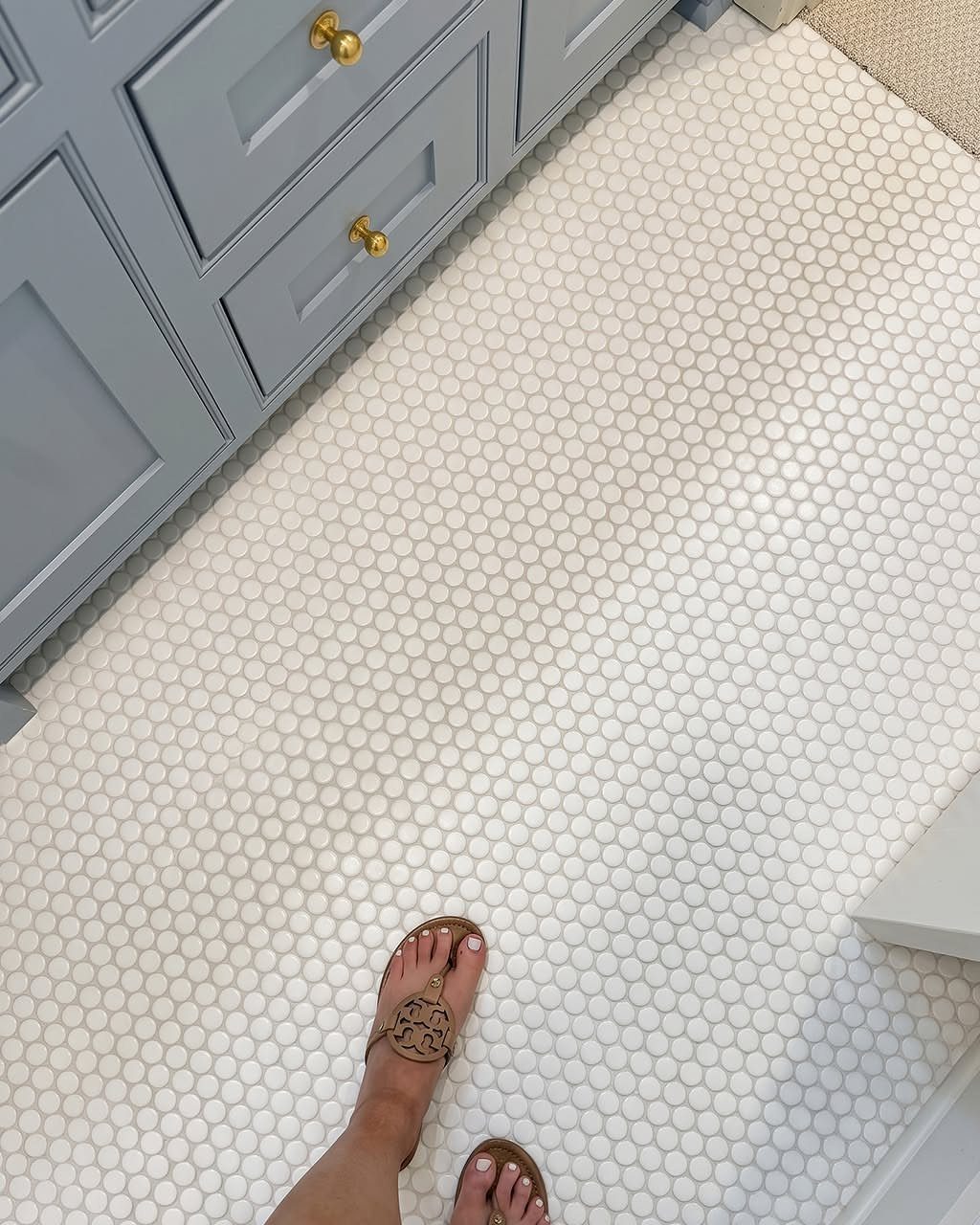 White penny tiles covering a bathroom floor next to light blue cabinets and a person's feet.